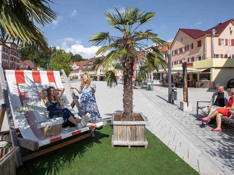 Vier Personen entspannen auf einer Bank und einem Liegestuhl auf dem Marktplatz.