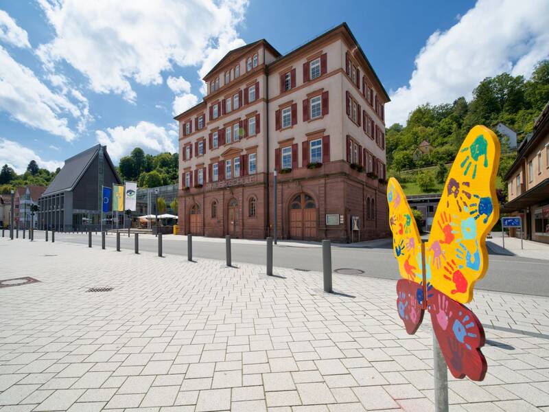Gelb-rot gestalteter Holzschmetterling auf dem Marktplatz mit der Musikschule und dem Rathaus im Hintergrund.