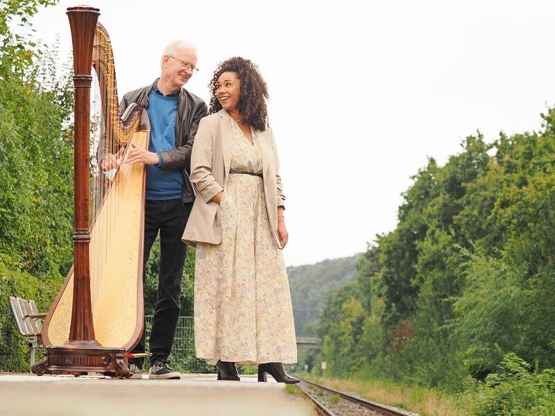 Hans Förster mit seiner Harfe und Kim Pauer in langem hellem Kleid stehen an einem Bahnsteig.