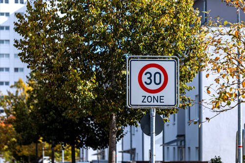 Ein Verkehrsschild, das auf den Beginn einer Tempo-30-Zone hinweist. Im Hintergrund befinden sich mehrere Bäume mit grünen Blättern und weiße Häuser.